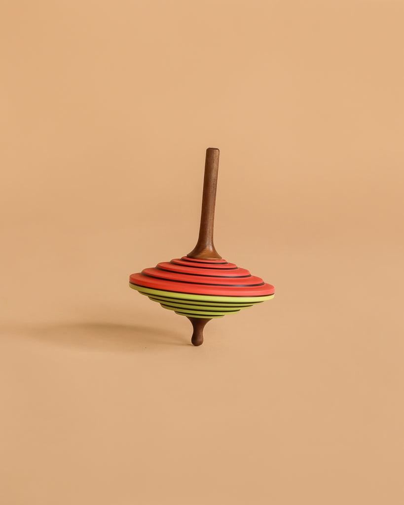 The Mader Tukan Spinning Top in green and red features vibrant, layered rings and stands upright on a beige background—a classic wooden toy inspired by Austrian craftsmanship.