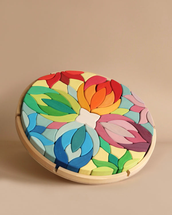 A round, colorful geometric puzzle featuring floral-like patterns with overlapping leaf shapes in various vibrant colors. The wooden Grimm's Lara Circle Flower Building Set challenges spatial thinking skills as you arrange them within a wooden frame, creating a visually appealing artwork against a plain beige background.