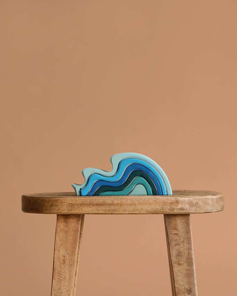 A wooden stool holds Grimm's Ocean Waves, Small—a blue and teal wave-shaped stacking toy for toddlers—set against a plain, light brown background.