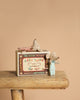 Two small Maileg mice from the Nursery Set with Brothers and Sisters rest in a matchbox bed with charming baby accessories, posed on a wooden stool against a beige background.