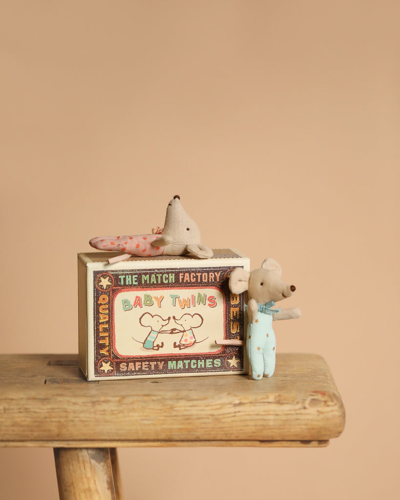 A small Maileg stuffed mouse in blue overalls stands beside a "Maileg Twin Baby Mice In Matchbox" box, illustrated with twin mice. Another mouse peers from the matchbox bed. Both sit on a rustic wooden stool against a beige background.