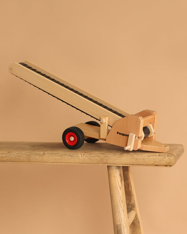 The Fagus Wooden Conveyor Belt, featuring "fagus" on its side and red and black wheels, sits angled upward on a rustic wooden bench against a beige background. It's part of the Fagus vehicles line.