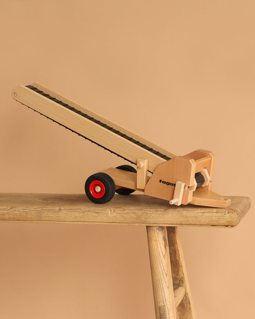 The Fagus Wooden Conveyor Belt, featuring "fagus" on its side and red and black wheels, sits angled upward on a rustic wooden bench against a beige background. It's part of the Fagus vehicles line.
