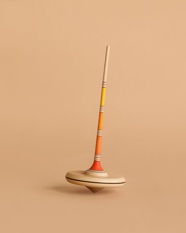 The Mader Arabella Spinning Top - Yellow features a long, slim handle and round base with orange, yellow, and beige stripes. It stands upright on a beige background and is crafted to meet strict toy safety standards.