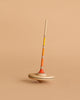 The Mader Arabella Spinning Top - Yellow features a long, slim handle and round base with orange, yellow, and beige stripes. It stands upright on a beige background and is crafted to meet strict toy safety standards.