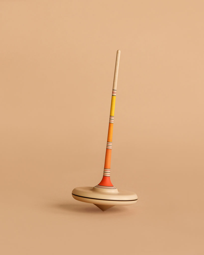The Mader Arabella Spinning Top - Yellow features a long, slim handle and round base with orange, yellow, and beige stripes. It stands upright on a beige background and is crafted to meet strict toy safety standards.