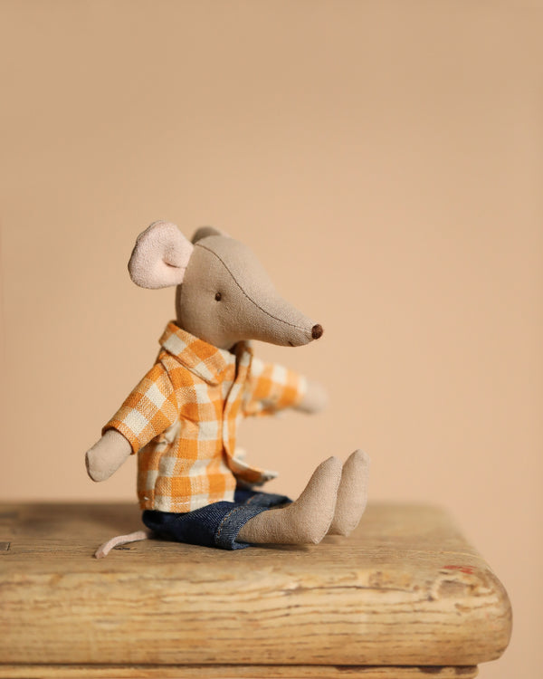 A small stuffed Dad mouse toy, named Maileg Dad Mouse With Yellow Shirt, dressed in a checked shirt and denim pants, sits on a wooden surface against a beige background.