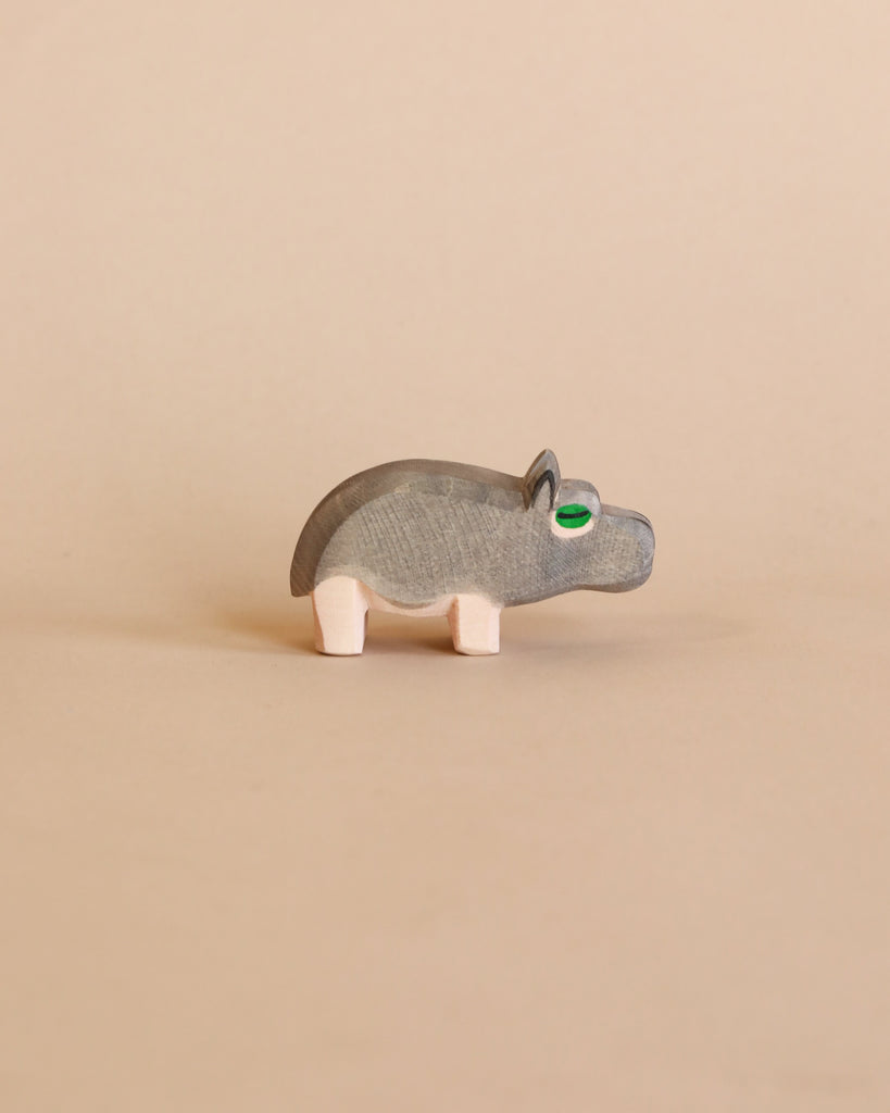 A small Ostheimer Hippopotamus figurine with visible wood grain, green eyes, and pale pink legs stands against a plain beige background.