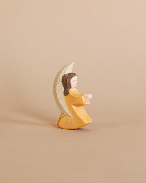 A small Ostheimer Little Angel - Orange figurine of a girl in a yellow dress, painted with a serene expression, embracing a white crescent moon, set against a plain beige background.