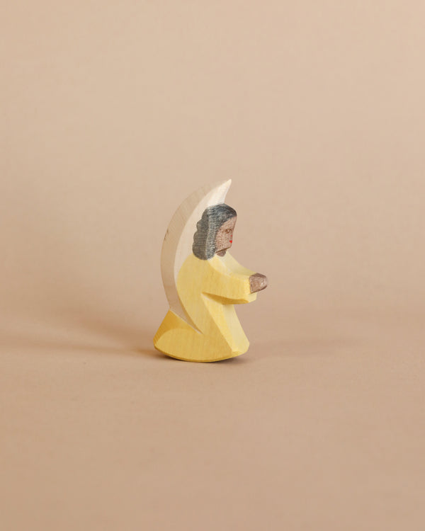 A handcrafted Ostheimer Little Angel - Yellow carved in the shape of a person wearing a yellow dress and a white headscarf, standing against a plain beige background.