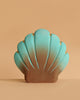 The Bumbu Handmade Wooden Big Shell with Pearl features a scallop shape, turquoise-to-brown gradient, and thin gold accents. It stands upright against a beige background, offering an elegant marine-inspired decorative piece.