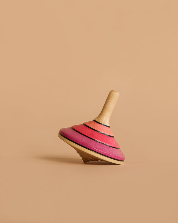 The Large Mader Tango Spinning Top - Pink, made of maple wood and adorned with pink and red stripes, rests on its side against a simple beige backdrop, showcasing its classic design and emphasis on toy safety.