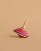 The Large Mader Tango Spinning Top - Pink, made of maple wood and adorned with pink and red stripes, rests on its side against a simple beige backdrop, showcasing its classic design and emphasis on toy safety.