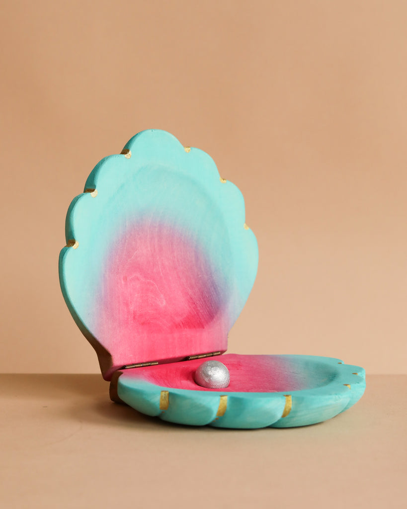 The Bumbu Handmade Wooden Big Shell with Pearl is a turquoise and pink, clam-shaped hinged box, open to display a single silver pearl inside. Handcrafted to evoke marine life, it is set against a plain light beige background.
