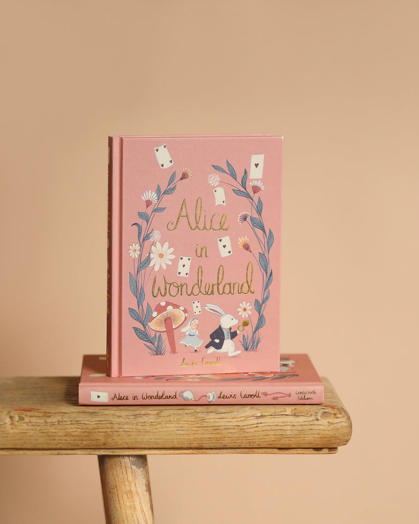 A pink hardcover Collector's Edition of "Alice in Wonderland" stands upright on a light brown wooden stool. Its cover, influenced by Lewis Carroll's story, depicts a rabbit, mushrooms, and playing cards within a floral wreath. Another book is positioned flat beneath it.