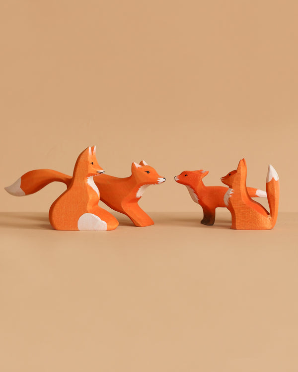 Four Bumbu Handmade Wooden Fox Family Set figurines, painted orange with white details, are arranged facing each other on a light tan background. The set includes two large and two small handmade wooden foxes.