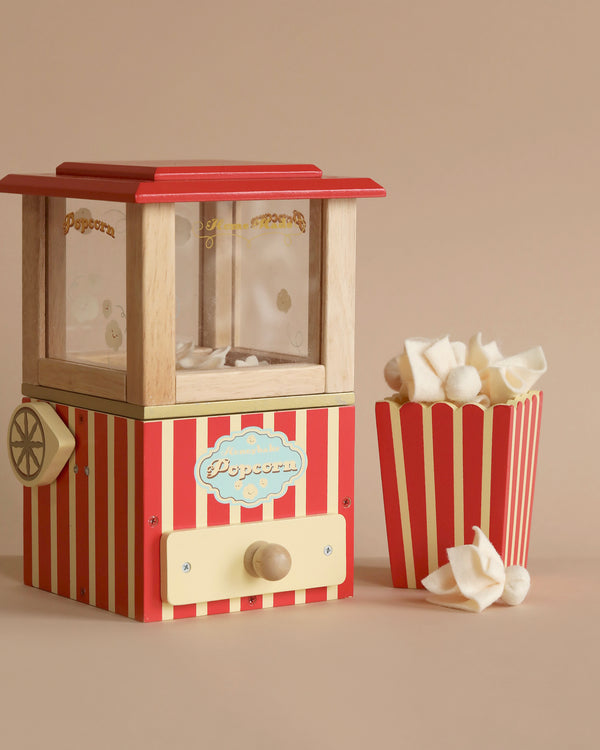 A small, Vintage Popcorn Maker with red and white stripes sits next to a matching popcorn container filled with popcorn. With its sustainable wood construction, the machine stands out against the plain beige background, highlighting its retro aesthetic and perfect for enhancing fine motor skills.