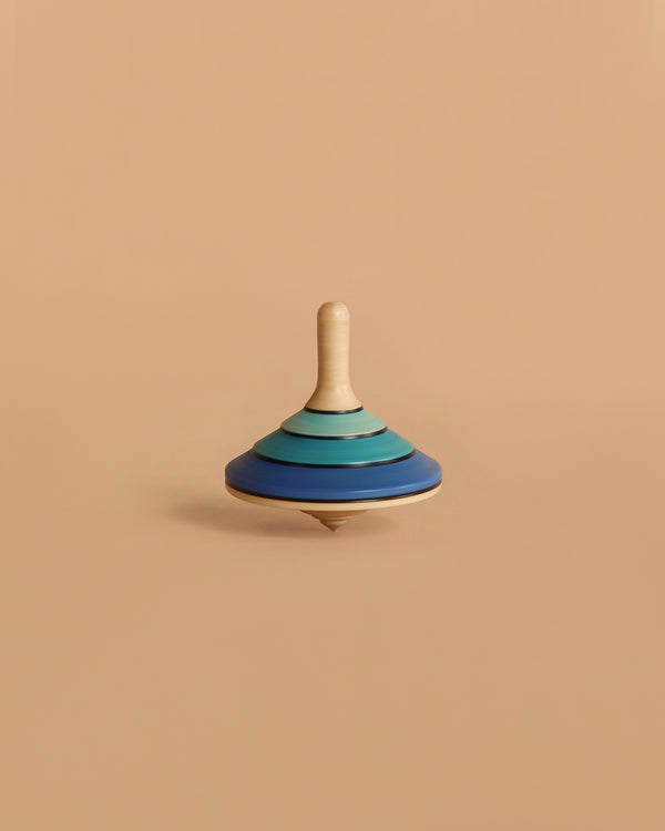 The Large Mader Tango Spinning Top - Dark Blue, crafted from maple wood with layered blue and teal rings, stands upright against a plain beige background, highlighting its safe design and elegant appearance.