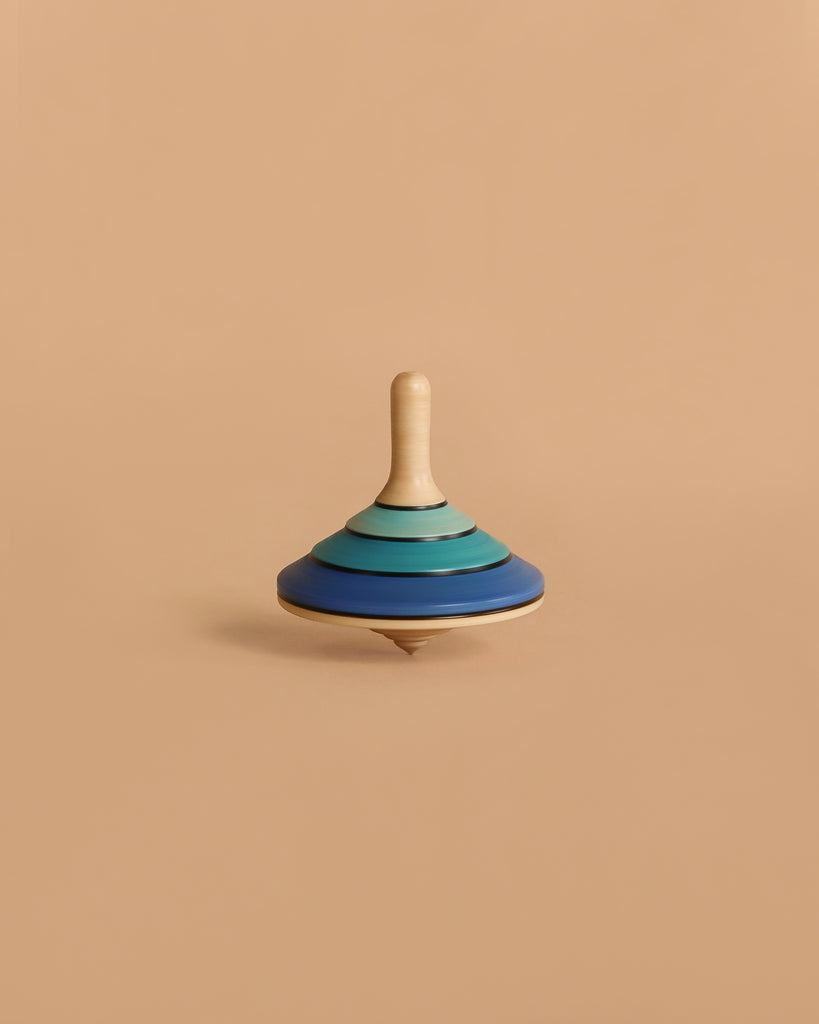 The Large Mader Tango Spinning Top - Dark Blue, crafted from maple wood with layered blue and teal rings, stands upright against a plain beige background, highlighting its safe design and elegant appearance.
