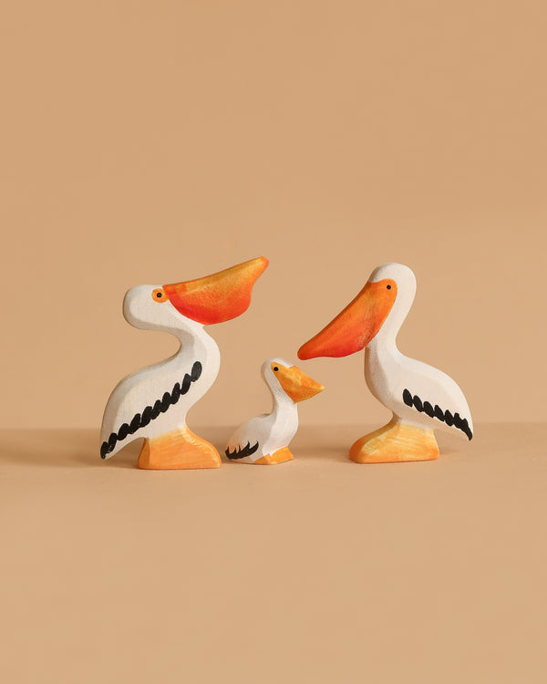 The Bumbu Handmade Wooden Pelicans Family Set features three sustainable wooden pelicans—two large and one small—painted white with orange beaks, feet, and black wings. Perfect for Montessori play and displayed against a beige background.