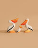 The Bumbu Handmade Wooden Pelicans Family Set features three sustainable wooden pelicans—two large and one small—painted white with orange beaks, feet, and black wings. Perfect for Montessori play and displayed against a beige background.