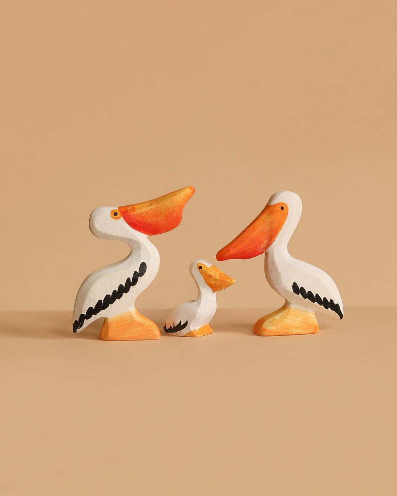 The Bumbu Handmade Wooden Pelicans Family Set features three sustainable wooden pelicans—two large and one small—painted white with orange beaks, feet, and black wings. Perfect for Montessori play and displayed against a beige background.