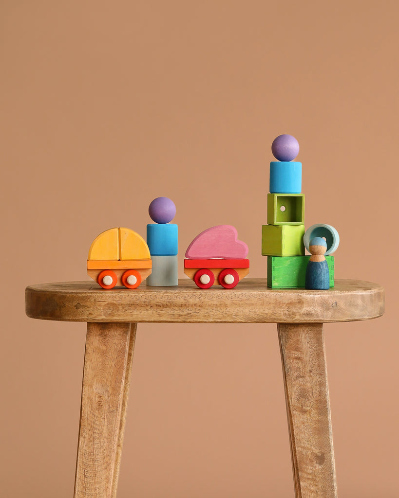 Grimm's Wooden Train Building Set displayed on a wooden table, featuring colorful blocks, cars, wagons, and a doll stacked playfully against a plain beige background.