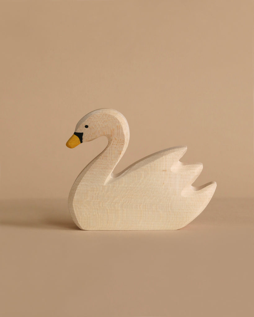 A small wooden toy crafted in the shape of a swan, painted in light beige with a yellow beak and a black mark near its eye, is displayed against a neutral beige background. This exquisitely handcrafted wood piece is reminiscent of the quality found in Holztiger Swan, Swimming figures.