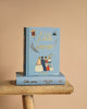 Two "Little Women" Collector's Edition hardcovers by Louisa May Alcott, with light blue covers, gold lettering, and illustrations of four women in vintage attire, rest on a rustic wooden stool against a beige background.