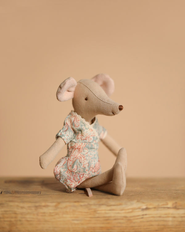 The Maileg Mum Mouse toy, with its large ears and pointed snout, wears a patterned dress while sitting on a wooden surface against a beige background, capturing a charming essence with its detailed design.