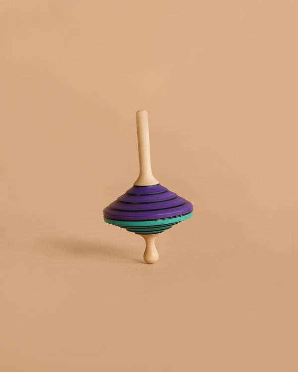The Mader Tukanino Spinning Top - Purple, crafted from maple with a cream handle and purple, black, and green stripes, stands upright against a beige background, capturing the playful spirit of Austrian toys.