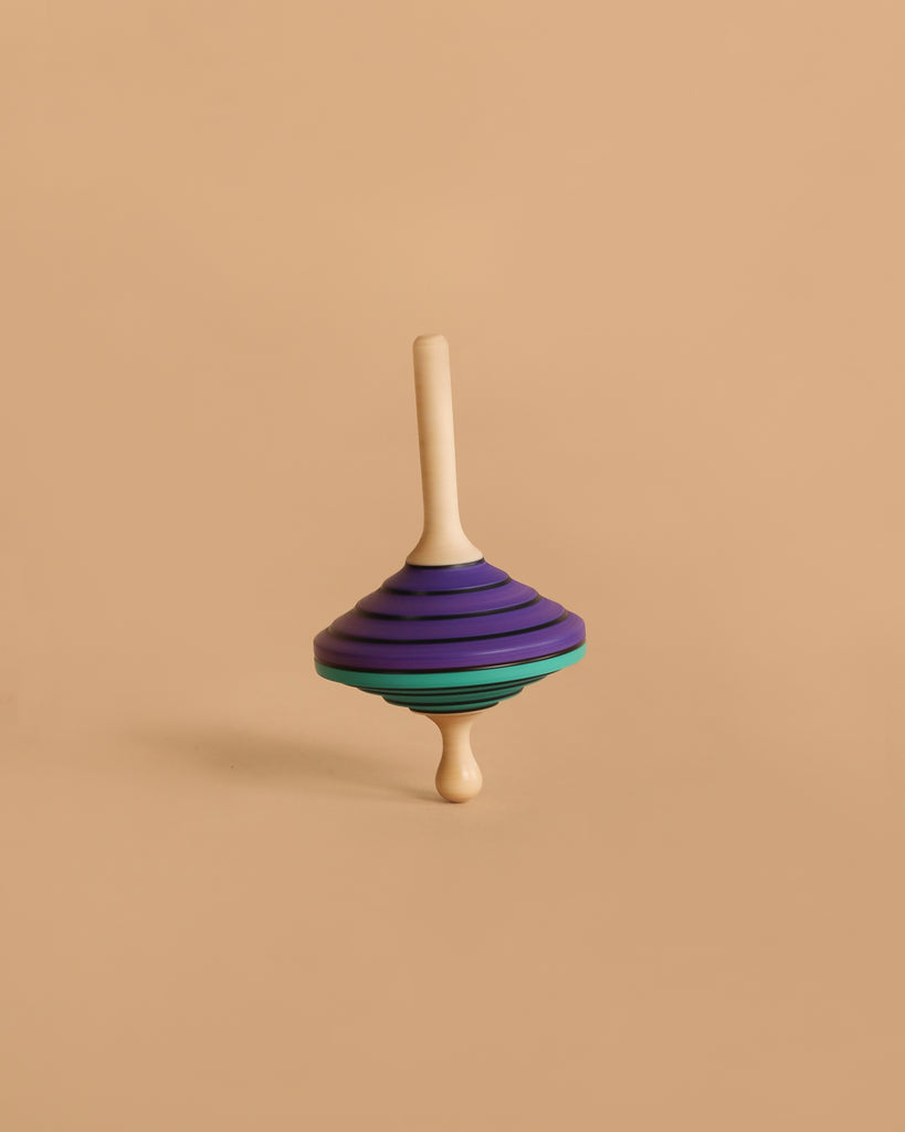The Mader Tukanino Spinning Top - Purple, crafted from maple with a cream handle and purple, black, and green stripes, stands upright against a beige background, capturing the playful spirit of Austrian toys.