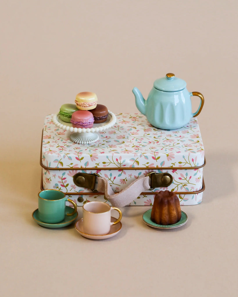 A small floral-patterned suitcase holds a teal teapot and colorful macarons. Below, the Maileg Afternoon Tea Set features two teacups with saucers and a small bundt cake, all against a beige background.