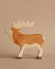 A handcrafted wooden toy moose with a simplified, cartoon-like design stands against a plain beige background. The Holztiger Stag is painted in warm shades of brown and cream, with dark brown hooves and a white face, featuring smooth curves and rounded edges.