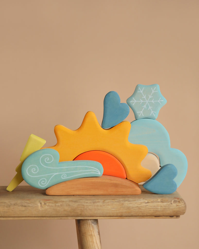 The Grimm's Weather Building Block Set features pastel-colored wooden pieces shaped like a sun, cloud, snowflake, and hearts in soft yellow, blue, and orange. These shapes are arranged on a wooden stool against a beige backdrop to spark young builders' imaginations.