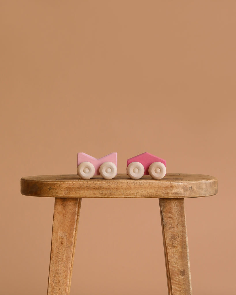 Two small Grimm's Stacking Cars - Pink with white wheels are placed side by side on a rustic wooden stool, set against a plain beige background.