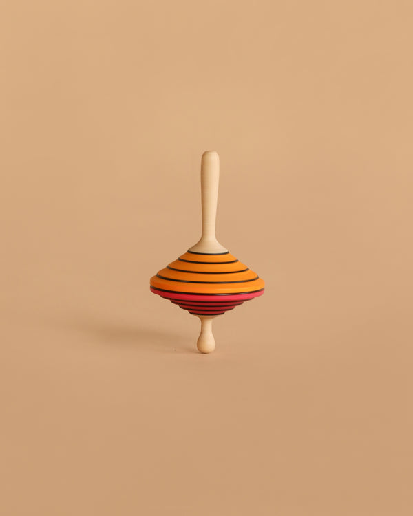 The Mader Tukanino Spinning Top - Orange, a maple wood fingertop with orange, yellow, green, and red stripes, stands upright on a plain beige background—a classic wooden toy from Austria.