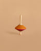 The Mader Tukanino Spinning Top - Orange, a maple wood fingertop with orange, yellow, green, and red stripes, stands upright on a plain beige background—a classic wooden toy from Austria.