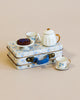A decorative Maileg Miniature Afternoon Treat Tea Set - Blue Madelaine arranged on a floral blue metal suitcase. The set includes a teapot, two cups, and a plate with a small tart, all on a light beige background.