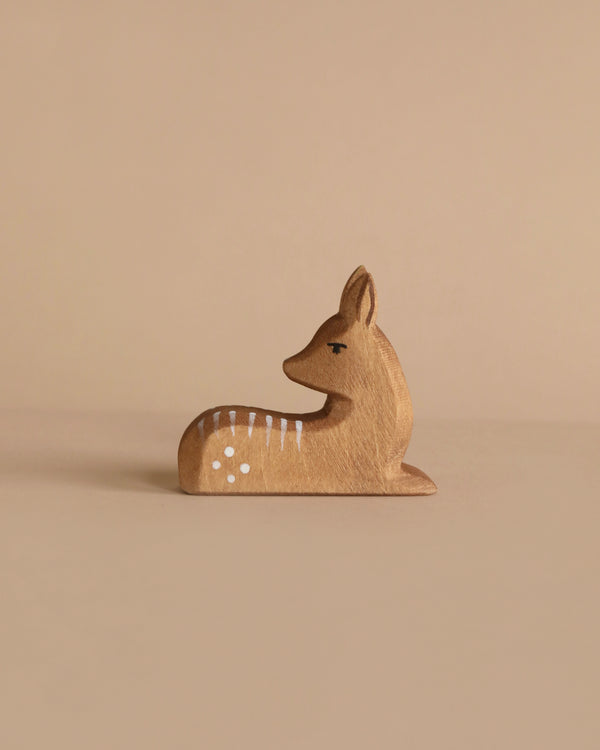 A small, light brown wooden figurine of a deer, the Ostheimer Fawn, Lying Down, rests on a neutral beige background. It is depicted in a lying down position, with its head upright and white spots along its back—perfect for imaginative play.