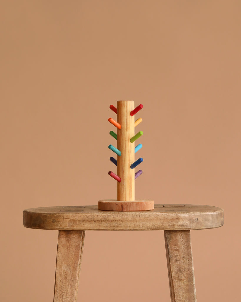 The Grimm's Sorting Helper for Building Rings - Rainbow, a small wooden tree-like stand with colorful pegs, is displayed on a rustic stool against a plain beige background—perfect for sorting games or ring storage.