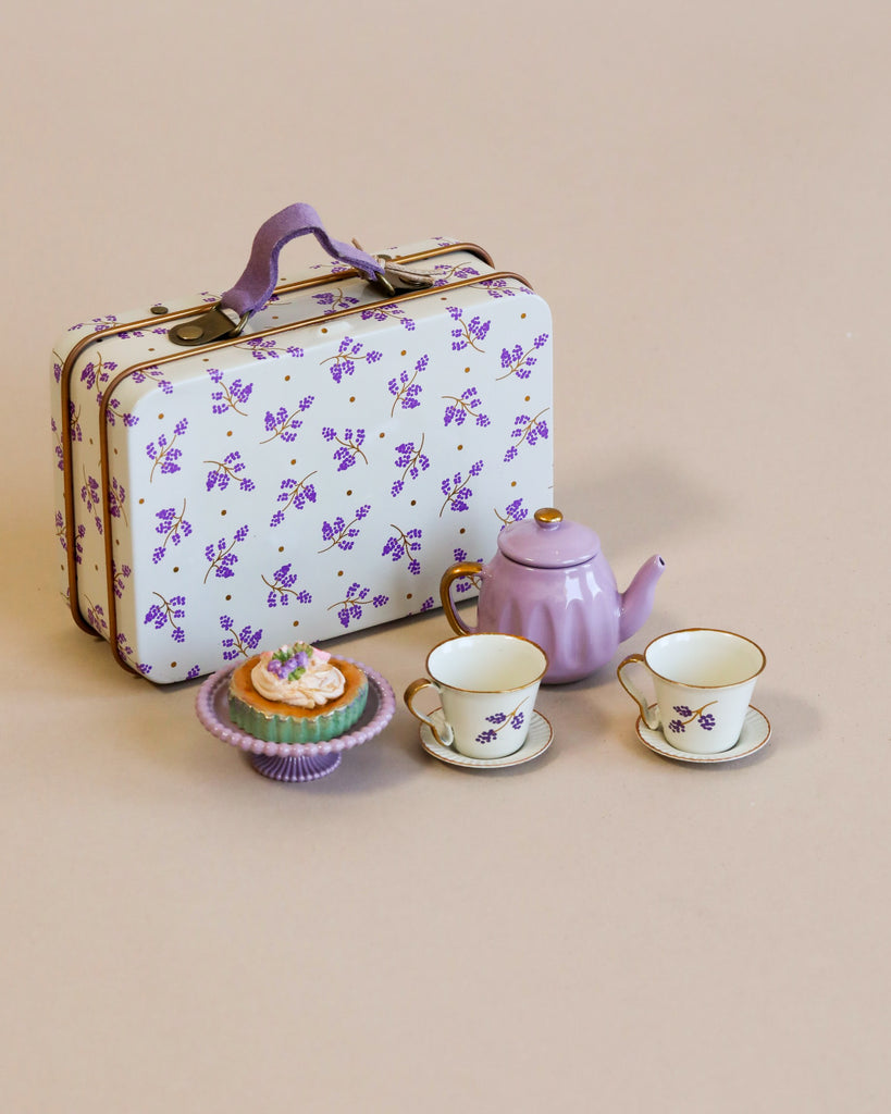 A Maileg Miniature Afternoon Treat Tea Set - Purple Madelaine with teapot, cups, and a cupcake beside a floral patterned suitcase on a plain beige background.