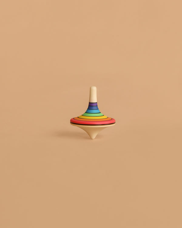 The Mader Rallye Rainbow Spinning Top, Large - Red features concentric rainbow colors on beech wood and stands upright on a plain light brown surface against a matching background.