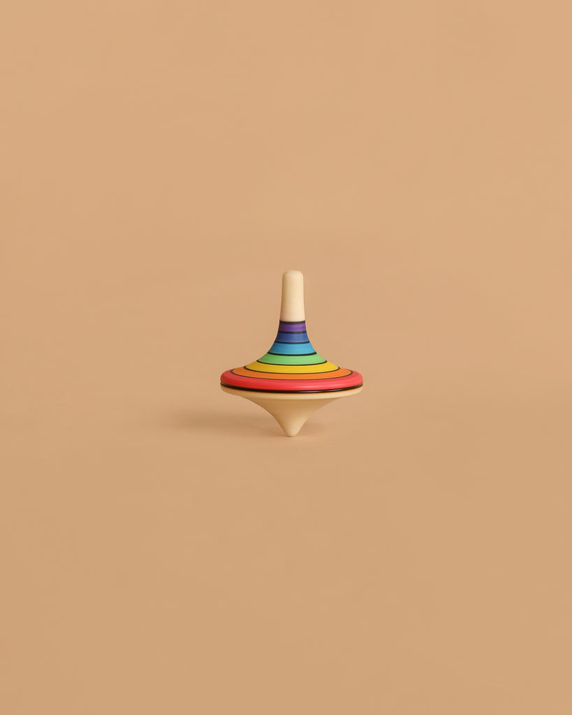 The Mader Rallye Rainbow Spinning Top, Large - Red features concentric rainbow colors on beech wood and stands upright on a plain light brown surface against a matching background.
