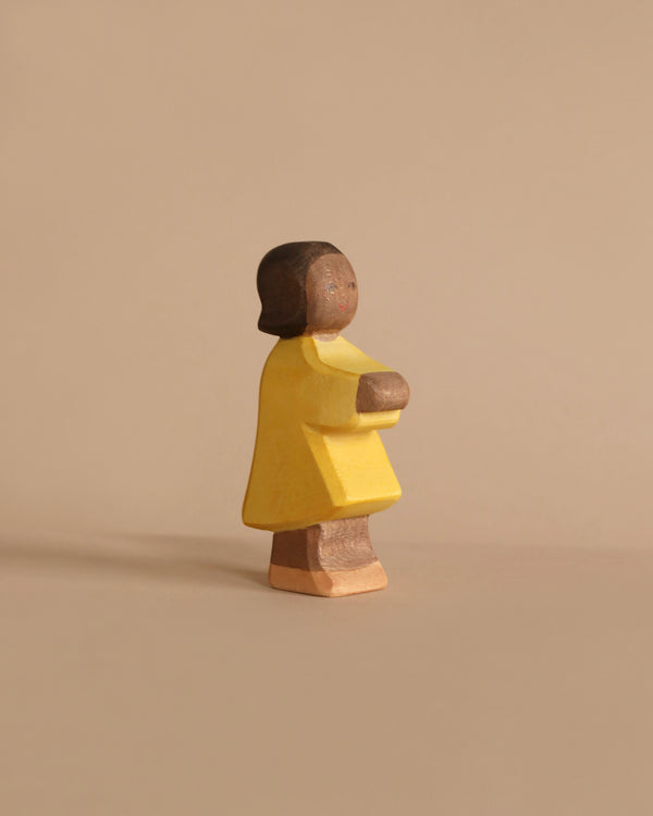 A wooden figurine of a person stands against a plain beige background, characteristic of the Ostheimer Daughter. The handcrafted wooden toy features a dark brown head and limbs, and it wears a yellow outfit. With its minimalist design and muted colors, it captures a side profile facing right, ideal for imaginative play.