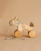 A Handmade Wooden Horse Pull Toy | Blue with four wheels, a piece of string attached at the front, and a painted heart and stripes on its body. The horse has a small, knotted tail and features a minimalist design. The background is a plain beige color.