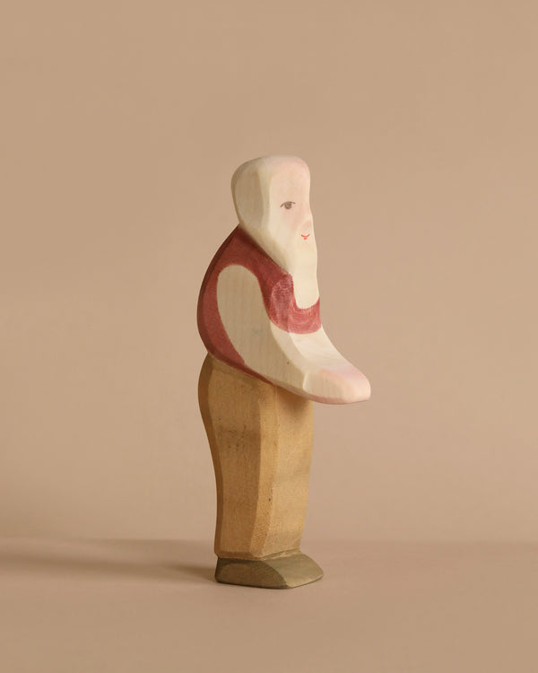 An Ostheimer Grandfather, a wooden figurine of a standing person with minimalistic features, handcrafted in the renowned Ostheimer tradition. The figure has a white head, a red shirt with a light pink arm, beige pants, and dark brown shoes against a plain beige background. The style is simplistic and blocky.