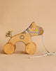 A Handmade Wooden Horse Pull Toy | Blue with painted details, a blue dotted harness, and a heart symbol on its side. This charming kids' toy with wheels features four circular wheels and a string attached to the front for pulling. The neutral beige background enhances its classic appeal.