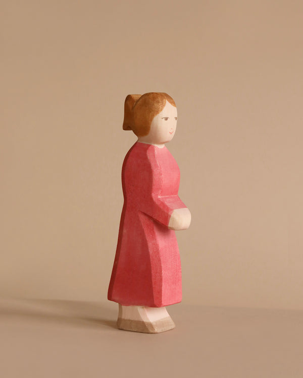 A small wooden figurine of a woman with light brown hair, wearing a long red dress. The Ostheimer Mother, Red Dress, is carved in a simplistic style and crafted from sustainably sourced materials. She faces to the right on a plain beige background.