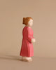 A small wooden figurine of a woman with light brown hair, wearing a long red dress. The Ostheimer Mother, Red Dress, is carved in a simplistic style and crafted from sustainably sourced materials. She faces to the right on a plain beige background.